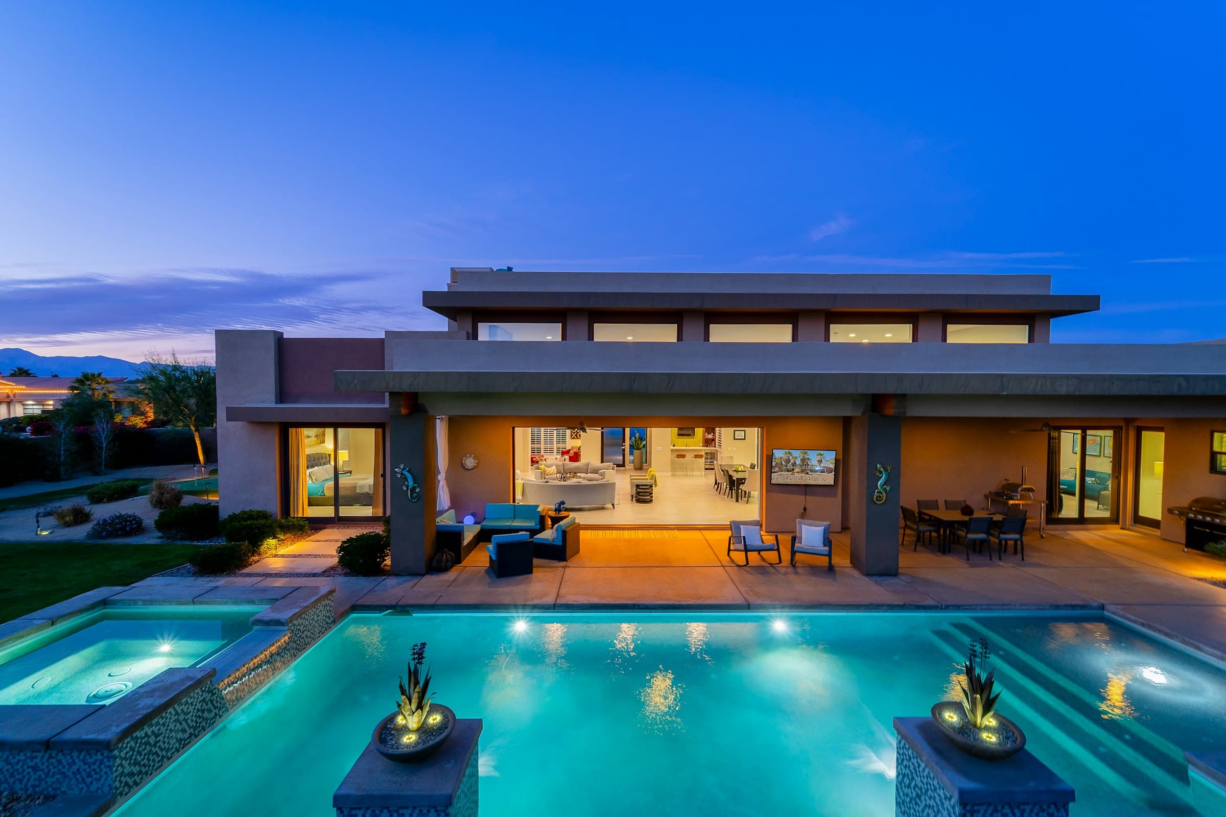 A backyard pool and spa lit bright blue at blue hour, fire bowls on the edge.