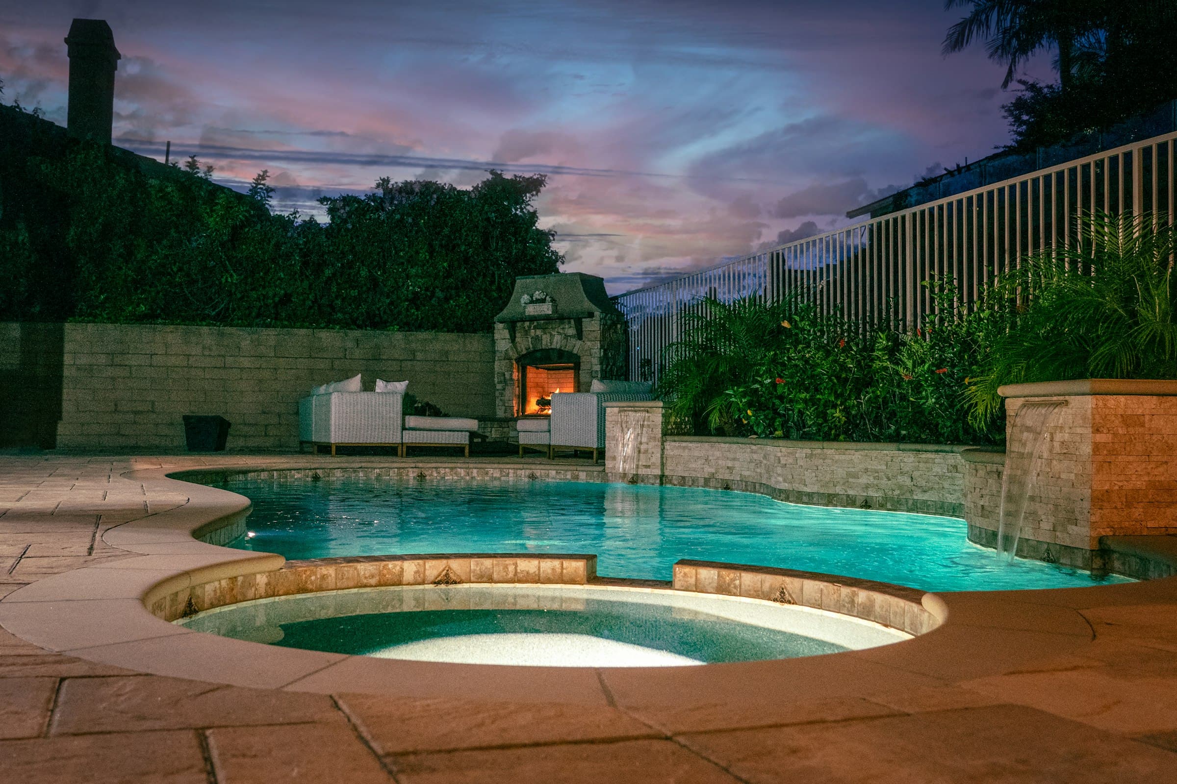 A pool and spa lit at dusk beside a stone fire feature and planting.