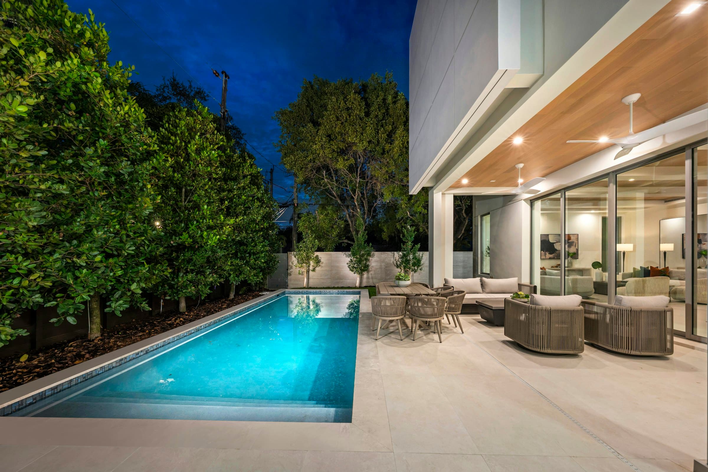 A lit pool beside a covered patio at dusk, a hedge along the side.